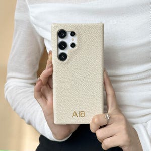 Könnte beinhalten: Cremefarbene Handyhülle mit einer strukturierten, lederartigen Oberfläche. Die rechteckige Hülle hat einen Kameraausschnitt und die goldenen Initialen "AIB". Die Hülle wird von einer Person gehalten.