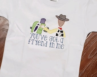 Pode incluir: Camiseta branca com uma imagem bordada de Woody e Buzz Lightyear de Toy Story. O texto "You've got a friend in me" está bordado abaixo da imagem.