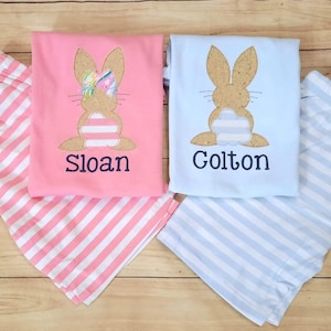 Może przedstawiać: Dwa komplety pasujących piżam dla dzieci. Jeden komplet jest r&oacute;żowy w białe paski i przedstawia brązowego kr&oacute;lika z r&oacute;żową kokardką. Drugi komplet jest niebieski w białe paski i przedstawia brązowego kr&oacute;lika. Piżamy są spersonalizowane z imionami "Sloan" i "Colton".