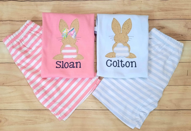 Może przedstawiać: Dwa komplety pasujących piżam dla dzieci. Jeden komplet jest r&oacute;żowy w białe paski i przedstawia brązowego kr&oacute;lika z r&oacute;żową kokardką. Drugi komplet jest niebieski w białe paski i przedstawia brązowego kr&oacute;lika. Piżamy są spersonalizowane z imionami "Sloan" i "Colton".