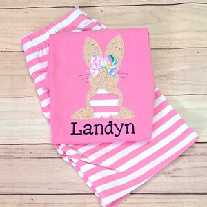 Może przedstawiać: R&oacute;żowe i białe pasiasty spodenki z r&oacute;żową koszulą z haftowanym kr&oacute;likiem z kokardką i imieniem "Landyn" w kolorze czarnym.