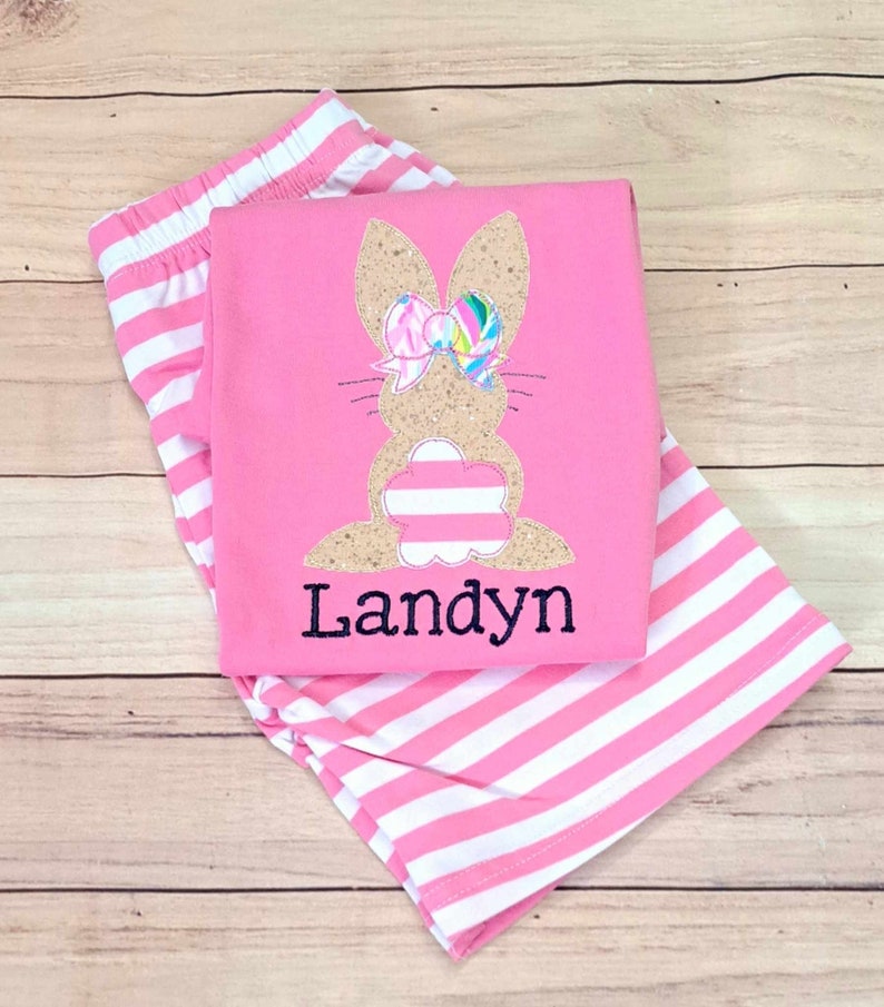 Może przedstawiać: R&oacute;żowe i białe pasiasty spodenki z r&oacute;żową koszulą z haftowanym kr&oacute;likiem z kokardką i imieniem "Landyn" w kolorze czarnym.