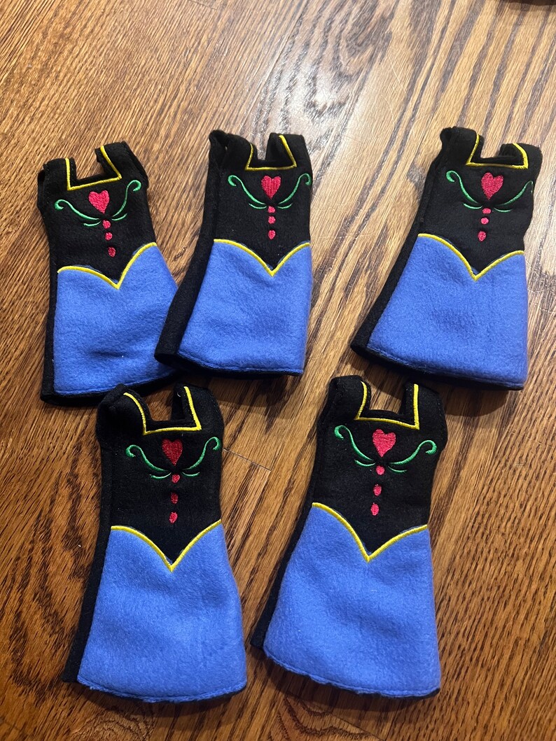 May include: Five felt doll dresses in blue, black, and yellow with red and green embroidery. The dresses feature a heart design and are perfect for a dollhouse or playset.