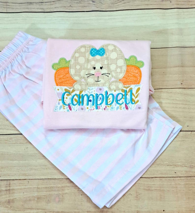May include: Pink and white striped shorts and a pink shirt with an embroidered bunny holding a carrot and a banner that says "Campbell".