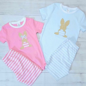 Może przedstawiać: Dwa komplety piżam dla dzieci, jeden r&oacute;żowy, a drugi niebieski, z aplikacją złotego błyszczącego kr&oacute;liczka z przodu. Piżamy mają białe paski na spodniach.