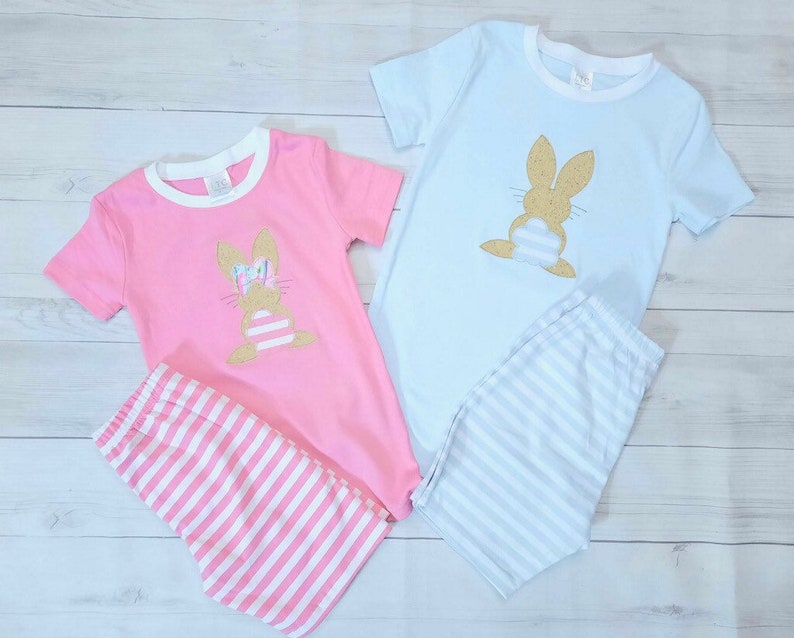 Może przedstawiać: Dwa komplety piżam dla dzieci, jeden r&oacute;żowy, a drugi niebieski, z aplikacją złotego błyszczącego kr&oacute;liczka z przodu. Piżamy mają białe paski na spodniach.