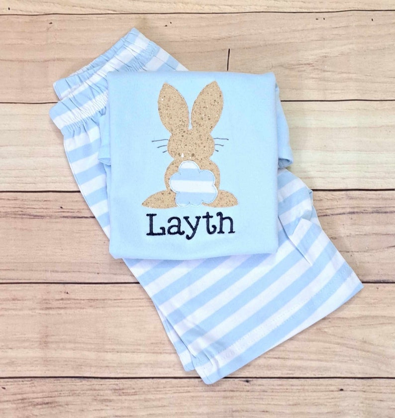 Może przedstawiać: Piżama w paski jasnoniebieskie i białe z jasnoniebieską koszulą z brązowym błyszczącym kr&oacute;likiem z niebiesko-białym paskowanym ogonem i imieniem "Layth" wyszytym z przodu.