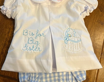 May include: White baby outfit with a Peter Pan collar and puff sleeves. The top has the embroidered text "B is for Big Sister" and a baby carriage design. The matching diaper cover is light blue and white gingham.