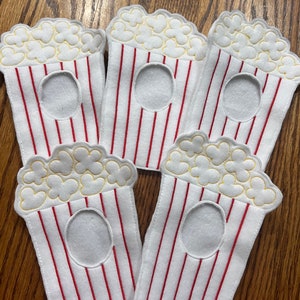 May include: Five white felt popcorn bags with red and white stripes. The bags have a yellow embroidered popcorn design and a hole cut out in the center.