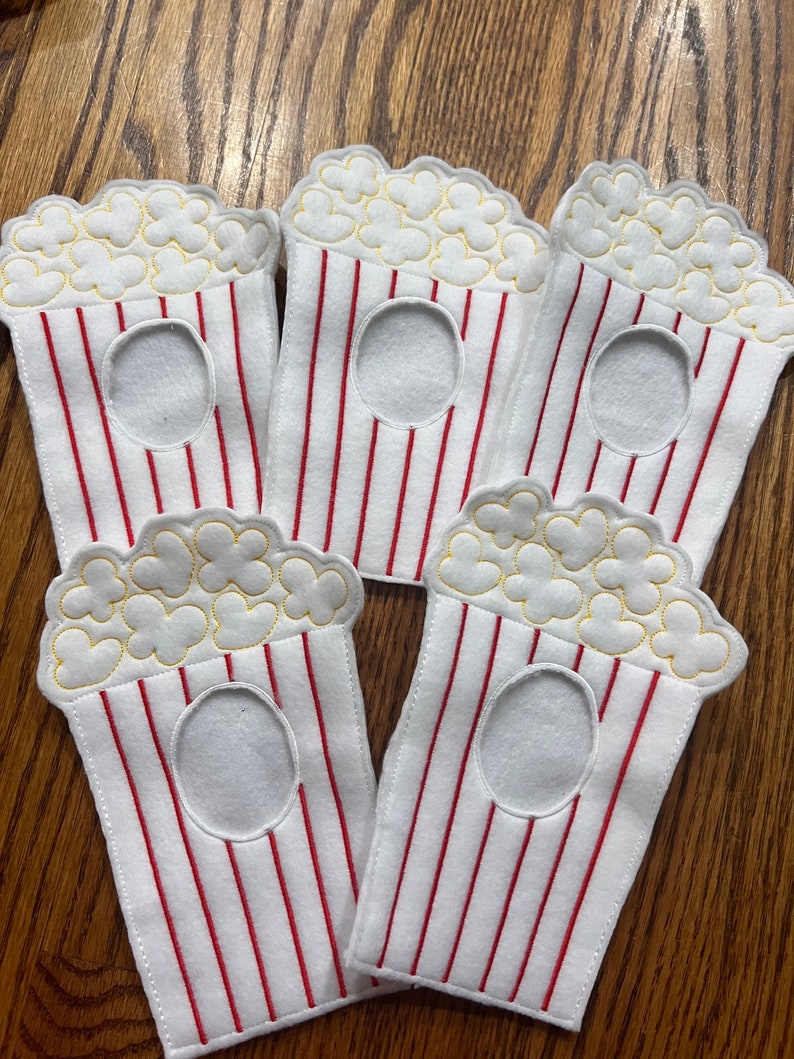 May include: Five white felt popcorn bags with red and white stripes. The bags have a yellow embroidered popcorn design and a hole cut out in the center.