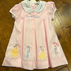 May include: A pink dress with white trim and embroidered Disney princesses. The dress has a Peter Pan collar and says "Once upon a time..." and "Campbell" on the front.