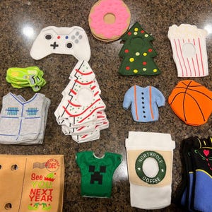 May include: A collection of felt Christmas ornaments, including a green tree, a red and white striped tree, a blue sweater, a basketball, a popcorn bucket, a donut, a video game controller, a green shirt with a Minecraft character, a brown box with the text "See You Next Year", and a coffee cup with the text "North Pole Coffee".