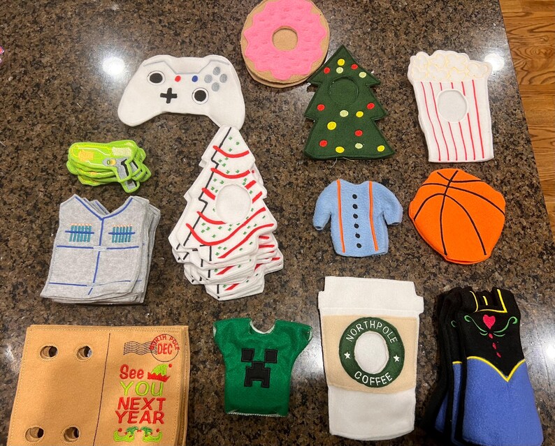 May include: A collection of felt Christmas ornaments, including a green tree, a red and white striped tree, a blue sweater, a basketball, a popcorn bucket, a donut, a video game controller, a green shirt with a Minecraft character, a brown box with the text "See You Next Year", and a coffee cup with the text "North Pole Coffee".