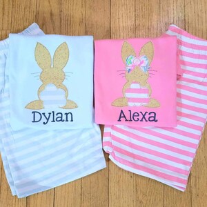 Może przedstawiać: Dwie pary paskowanych piżam z aplikacjami kr&oacute;liczk&oacute;w. Niebieska piżama ma kr&oacute;liczka z brokatem w kolorze złotym z wyhaftowanym imieniem "Dylan" poniżej. R&oacute;żowa piżama ma kr&oacute;liczka z brokatem w kolorze złotym z r&oacute;żową kokardką i wyhaftowanym imieniem "Alexa" poniżej.