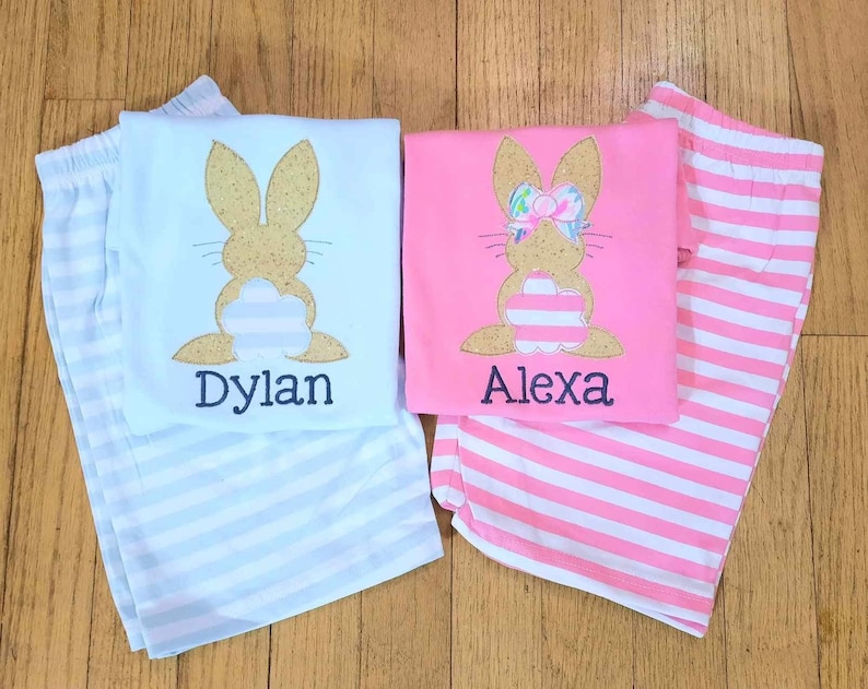 Może przedstawiać: Dwie pary paskowanych piżam z aplikacjami kr&oacute;liczk&oacute;w. Niebieska piżama ma kr&oacute;liczka z brokatem w kolorze złotym z wyhaftowanym imieniem "Dylan" poniżej. R&oacute;żowa piżama ma kr&oacute;liczka z brokatem w kolorze złotym z r&oacute;żową kokardką i wyhaftowanym imieniem "Alexa" poniżej.