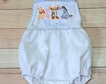Könnte beinhalten: Weißer Baby Strampler mit blau-weiß gestreiften Trägern. Der Strampler ist mit Winnie Puuh Figuren und dem Namen "Charlee" auf der Vorderseite bestickt.