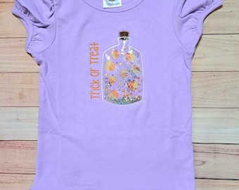 May include: Purple short-sleeved shirt with a ruffle hem. The shirt features an embroidered design of a glass jar filled with candy corn and the text "Trick or Treat".