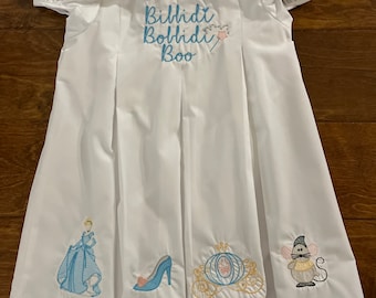 Op de afbeelding: Witte jurk met blauwe borduursels met de tekst "Bibbidi Bobbidi Boo" en afbeeldingen van Assepoester, een glazen muiltje en een koets.