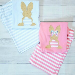 Może przedstawiać: Dwie pary paskowanych piżam z aplikacją kr&oacute;lika. Jedna para jest jasnoniebieska z brązowym kr&oacute;likiem, a druga jest r&oacute;żowa z brązowym kr&oacute;likiem z kokardką.