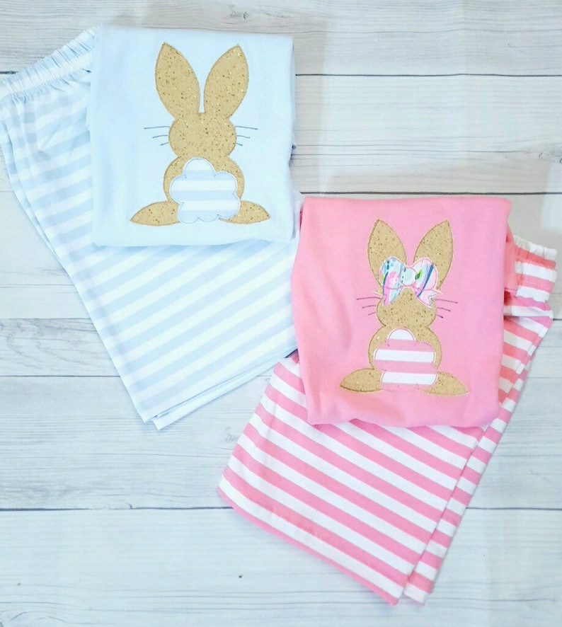 Może przedstawiać: Dwie pary paskowanych piżam z aplikacją kr&oacute;lika. Jedna para jest jasnoniebieska z brązowym kr&oacute;likiem, a druga jest r&oacute;żowa z brązowym kr&oacute;likiem z kokardką.