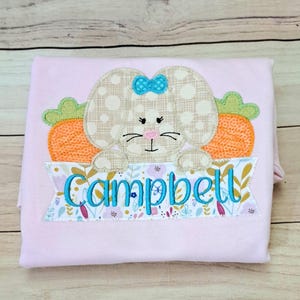May include: Pink personalized Easter shirt with a brown bunny holding a carrot and a banner with the name "Campbell" embroidered on it.