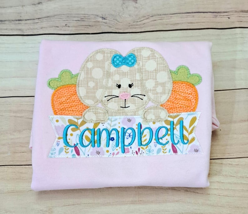 May include: Pink personalized Easter shirt with a brown bunny holding a carrot and a banner with the name "Campbell" embroidered on it.