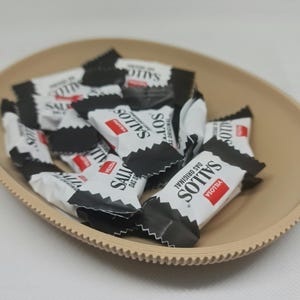 Könnte beinhalten: Eine beige ovale Servierschale gefüllt mit einzeln verpackten Bonbons. Die Süßigkeitenverpackungen sind weiß mit schwarzen Akzenten und rotem Text mit der Aufschrift "Sallos Das Original". Die Schale hat einen strukturierten, geriffelten Rand.