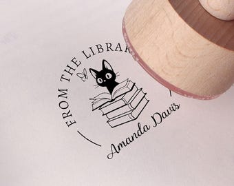 May include: A round wooden rubber stamp pressing onto a book page, leaving a black ink impression of a cat sitting on a stack of books with the text FROM THE LIBRARY and the name Amanda Davis printed in a circular design.