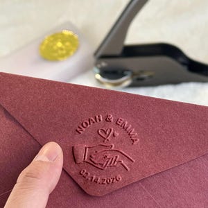 Könnte beinhalten: Ein weinroter Umschlag mit einem geprägten Design mit den Namen "Noah & Emma", einem Herzen, Händen, die sich halten, und dem Datum "02.14.2026". Ein goldener Siegel und eine schwarze Siegelpresse befinden sich im Hintergrund. Der Umschlag wird von einer Hand gehalten.