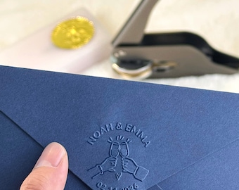 Könnte beinhalten: Ein marineblauer Umschlag mit geprägtem Text "NOAH & EMMA" über zwei anstoßenden Sektgläsern und dem Datum "02.14.2026". Ein goldener Siegel und eine silberne Siegelpresse befinden sich im Hintergrund.