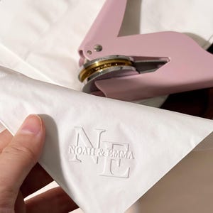 Puede incluir: Un sello de relieve rosa claro con una matriz dorada y plateada, junto a un papel blanco con el texto en relieve "NOAH & EMMA". El sello está diseñado para crear impresiones personalizadas en papel.