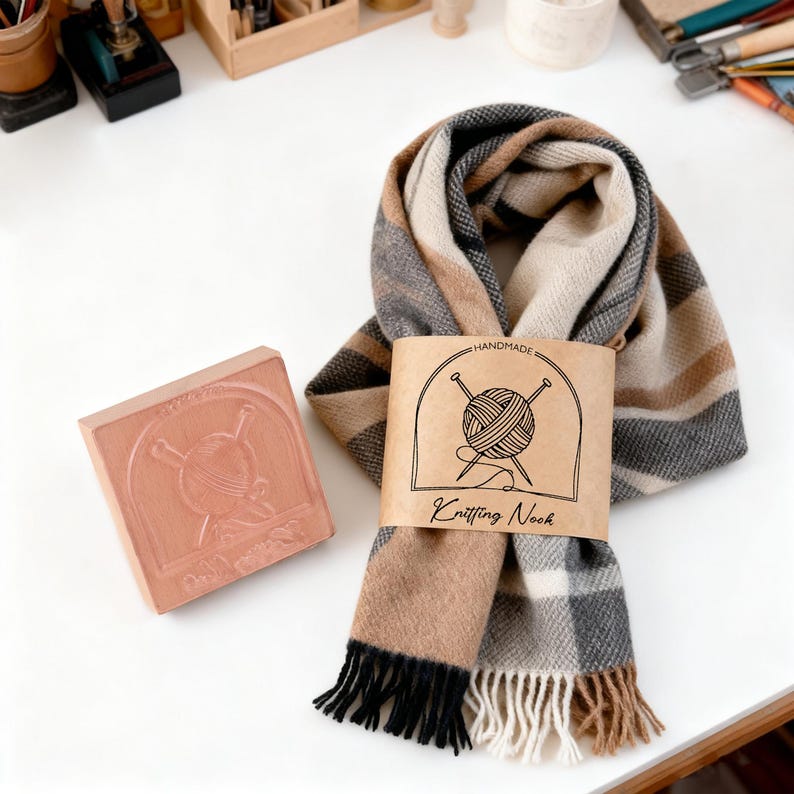 Pu&ograve; includere: Sciarpa fatta a mano con motivo a quadri marrone, beige e grigio, con frange nere e beige. La sciarpa &egrave; avvolta con un'etichetta che recita "Knitting Nook" e un logo di filato e aghi. &Egrave; presente anche un timbro quadrato con un disegno a maglia.
