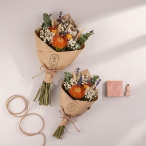 Pu&ograve; includere: Due bouquet di fiori secchi avvolti in carta marrone, legati con spago. I bouquet presentano protea arancioni, gipsofila bianca ed eucalipto verde. Sono presenti anche una saponetta e un flaconcino.