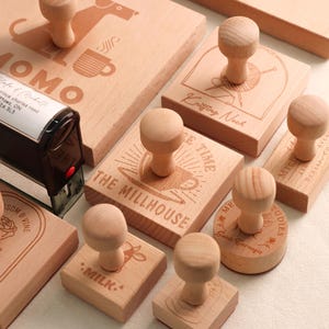 May include: A selection of wooden stamps with different designs and text. The stamps include images of a dog, a coffee cup, a ball of yarn, and the words "THE MILLHOUSE" and "MILK." Each stamp has a wooden handle.