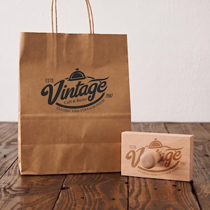 Puede incluir: Una bolsa de papel marrón con asas de papel trenzado y un logotipo impreso en negro que dice ESTD. Vintage Café & Resto 1987 CLASSIC AND VINTAGE FOOD, acompañada de un sello de goma de madera rectangular con el mismo diseño de logotipo.