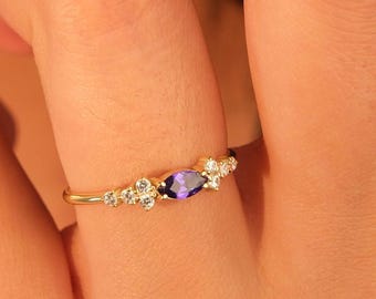 May include: A gold ring featuring a marquise-cut purple gemstone, flanked by small, round, clear gemstones. The ring has a delicate band and is set against a skin-toned background.