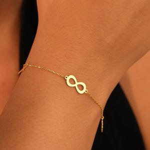 May include: A delicate gold-colored bracelet featuring an infinity symbol as its central charm. The bracelet is worn on a wrist, with the chain and charm clearly visible against the skin. The bracelet is a simple, elegant piece of jewelry.