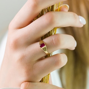 Peut inclure: Une bague en or avec une pierre carrée rouge. La bague est sur le doigt d'une personne.
