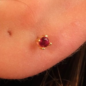 Może przedstawiać: Złota kolczyka sztyftowa z okrągłym, głębokim czerwonym kamieniem szlachetnym. Kolczyk osadzony jest w czteroramiennej oprawie, podkreślającej żywy kolor kamienia. Kolczyk jest mały i delikatny.