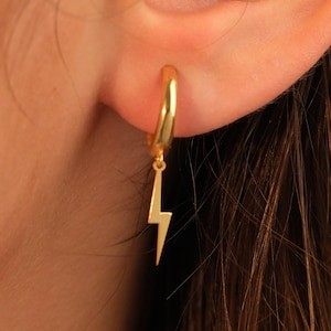 Op de afbeelding: Een goudkleurige oorring met een bliksemcharm. De oorring heeft een eenvoudig, elegant ontwerp met een gepolijste afwerking. De bliksemcharm hangt aan de onderkant van de ring.