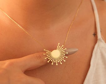 May include: A gold-colored necklace featuring a sunburst pendant. The pendant has a circular center with radiating lines ending in small, round accents. The necklace is worn on a person's chest, with the chain visible.