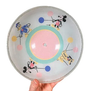 Może przedstawiać: Przezroczysty, okrągły, zabytkowy klosz do lampy sufitowej z postaciami Disneya, Myszki Miki i Kaczora Donalda. Lampa ma różową, niebieską i żółtą kolorystykę z falistą krawędzią. Klosz jest trzymany przez dłoń.