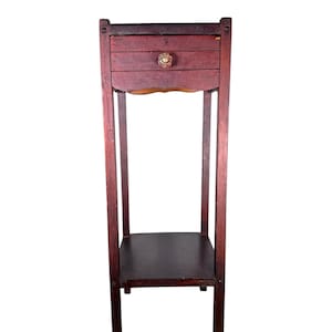 May include: A tall, dark reddish-brown wooden plant stand featuring a single drawer with a small, clear, faceted glass knob. The stand has four straight legs and a lower square shelf positioned between the legs, providing a stable base.