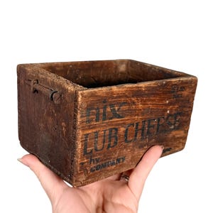 May include: A small, weathered wooden box with the text "Nix Lub Cheese" printed in black. The box is rectangular with a square opening and appears to be an antique. The box is held by a hand.