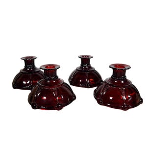 Op de afbeelding: Vier robijnrode glazen kandelaars. Elke houder heeft een uitlopende basis, een bolvormig middengedeelte en een kleine, cirkelvormige bovenkant. Het glas heeft een glanzende afwerking en een decoratief patroon rond de basis.