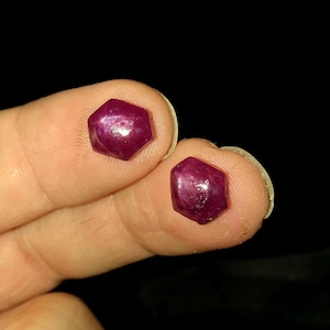 May include: Two loose, deep magenta-colored gemstones with a distinct hexagonal shape. Each stone features a faceted surface that reflects light, showcasing a rich, saturated red-purple hue and a smooth, polished appearance throughout the structure.