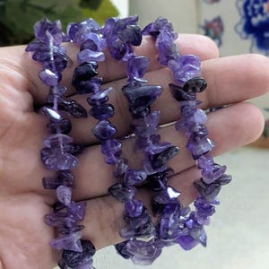 May include: Three strands of amethyst chip bead bracelets. The beads are a mix of light and dark purple, strung together. The bracelets are held in a hand, showcasing the natural color and texture of the gemstones.