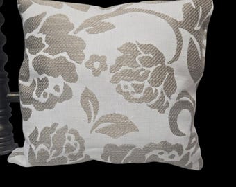 May include: Square decorative pillow with a white background and a large floral pattern in shades of beige and gray. The pillow is made of a textured fabric and is likely used for home decor.