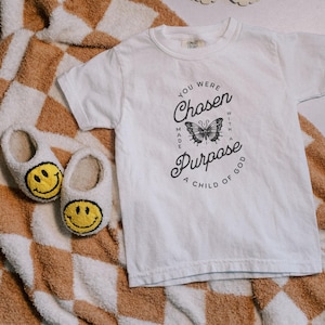 Puede incluir: Camiseta blanca con un diseño gráfico negro que presenta una mariposa y el texto "You were chosen with a purpose, a child of God".