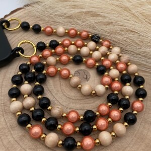 Könnte beinhalten: Ein mehrsträngiger Handy-Anhänger mit schwarzen, beigefarbenen und pfirsichfarbenen Perlen. Die Perlen sind auf einer goldenen Kette mit einem goldenen Verschluss aufgefädelt.